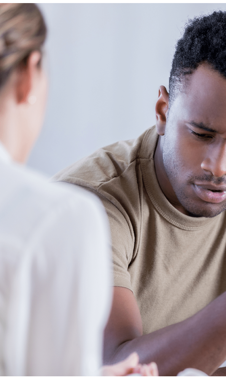 In this closeup, a young man wearing a military issue t-shirt and holding a camouflage cap looks down with a pained expression. He is sharing his problems with an unrecognizable counselor.