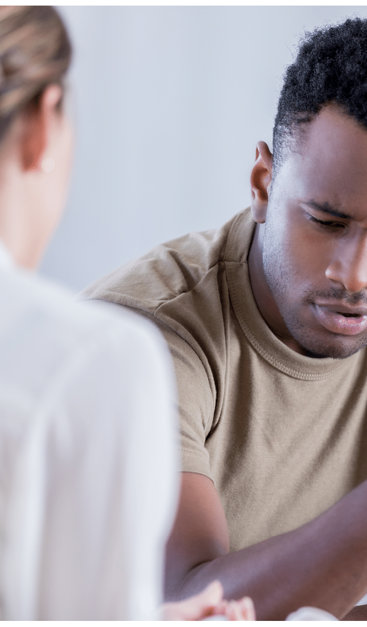 In this closeup, a young man wearing a military issue t-shirt and holding a camouflage cap looks down with a pained expression. He is sharing his problems with an unrecognizable counselor.