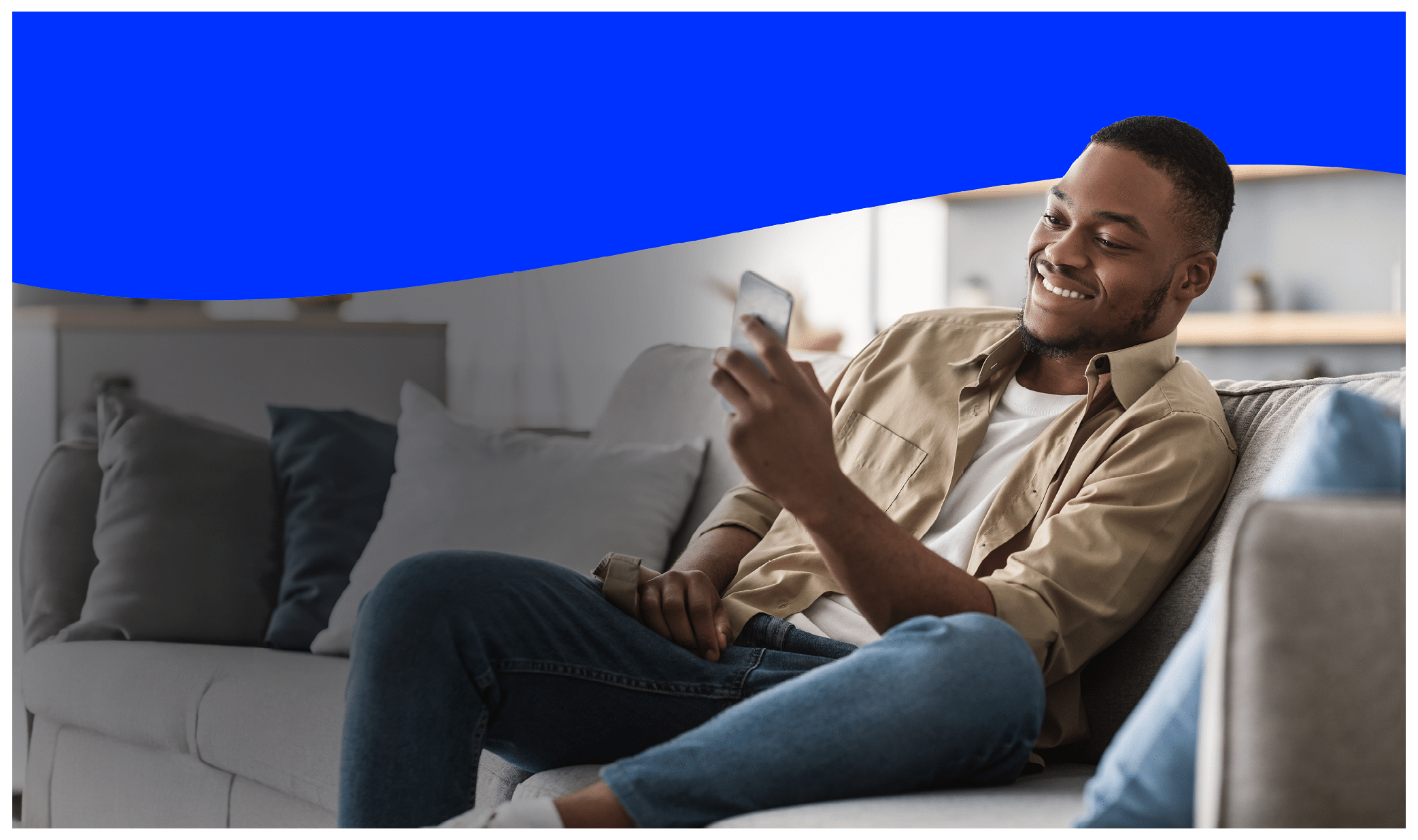 Side View Of African American Guy Using Smartphone Browsing Internet Sitting On Couch At Home. Black Man Texting On Cellphone Or Using New Application On Mobile Phone Indoors