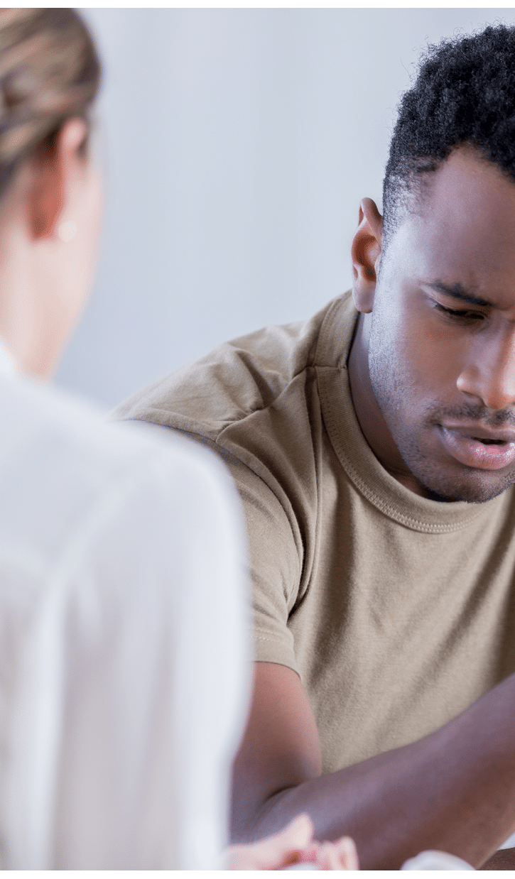 In this closeup, a young man wearing a military issue t-shirt and holding a camouflage cap looks down with a pained expression. He is sharing his problems with an unrecognizable counselor.