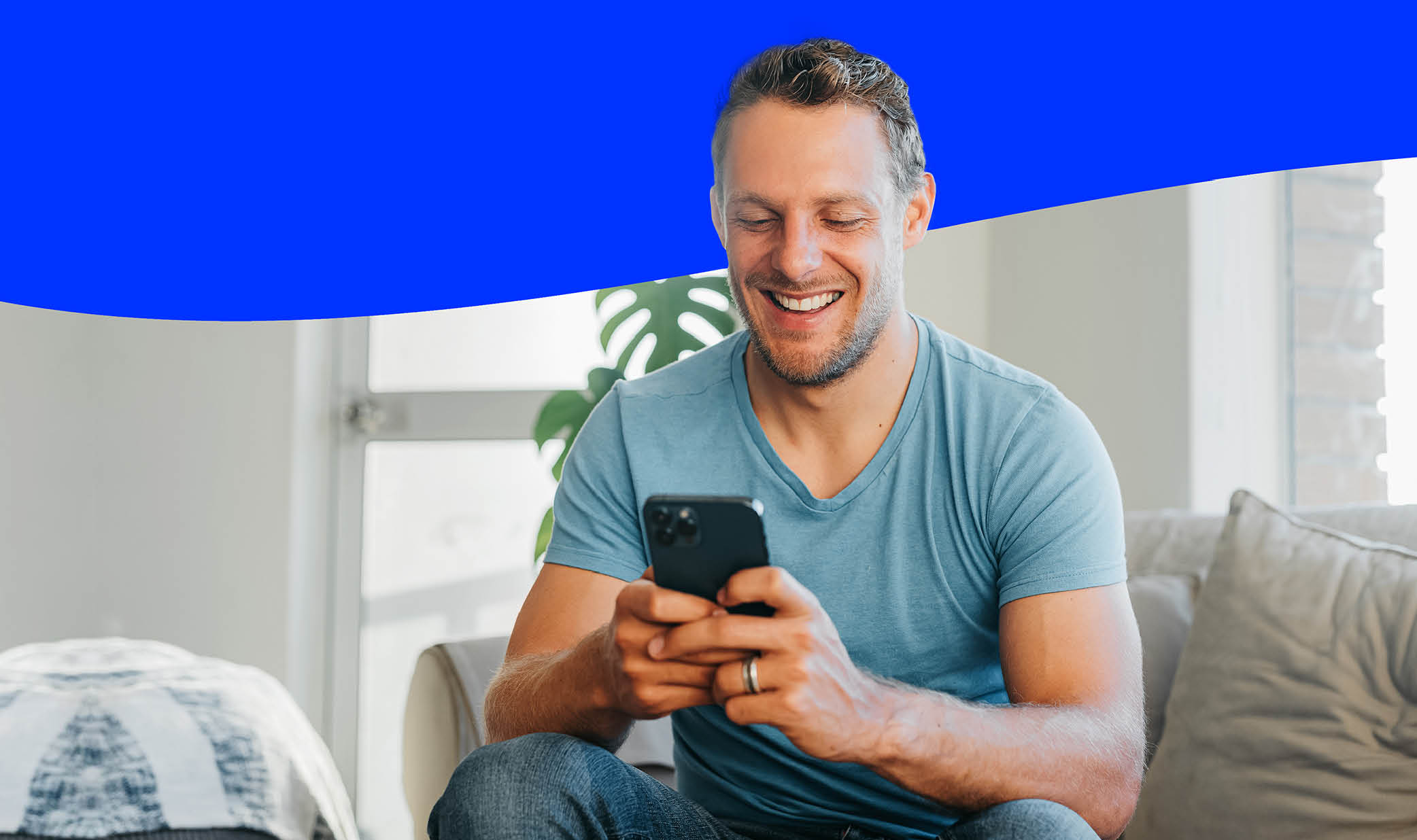 Side View Of African American Guy Using Smartphone Browsing Internet Sitting On Couch At Home. Black Man Texting On Cellphone Or Using New Application On Mobile Phone Indoors