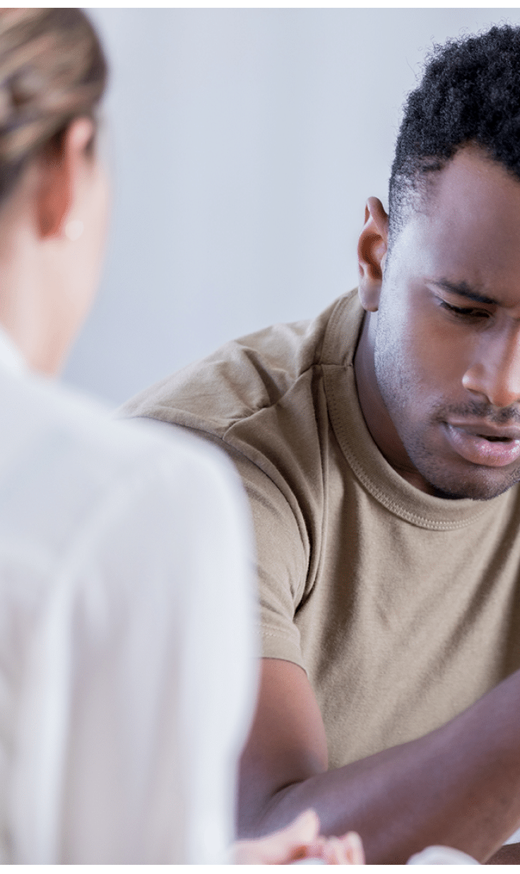 In this closeup, a young man wearing a military issue t-shirt and holding a camouflage cap looks down with a pained expression. He is sharing his problems with an unrecognizable counselor.